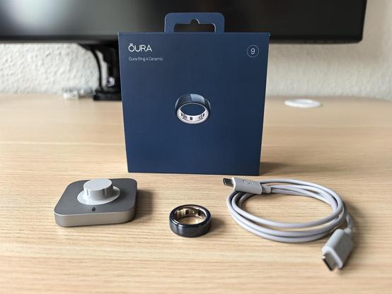 Oura Ring 4 Ceramic in Midnight (Dunkelblau) samt Zubehör und Verpackung auf einem Schreibtisch.