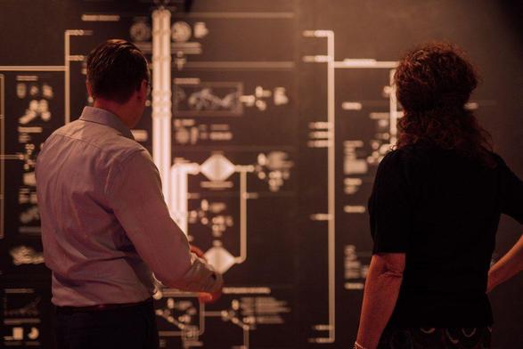 Twee mensen kijken naar "Anatomy of an AI system", een visuele weergave van hoe AI systemen aan elkaar gelinkt zijn, met zilveren lijnen en woorden tegen een zwarte achtergrond.