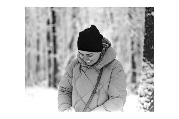 A black and white film photograph of a woman standing in a snowy forest. She is wearing a puffer coat and a beanie, looking down with a bright, happy smile.