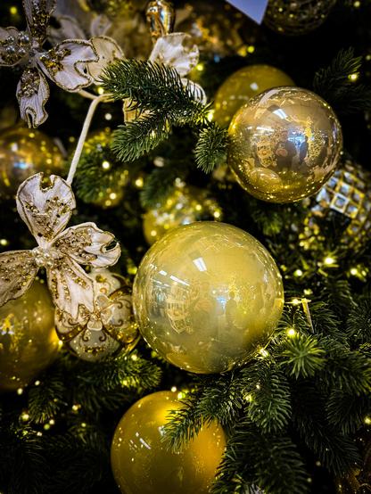 The image shows a close-up view of a decorated evergreen tree branch, likely part of a Christmas tree. Several golden ornaments of varying sizes are prominently displayed among the dark green pine needles. There are also decorative golden bows and embellishments scattered throughout the branch. The ornaments have a glossy, reflective surface and appear to be spherical in shape. The overall lighting is dim, creating highlights on the ornaments and a sense of depth within the branches.

Provided by @altbot, generated privately and locally using Gemma3:27b