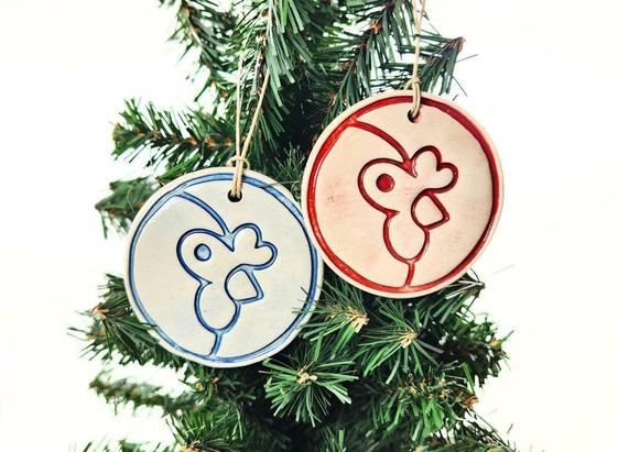 Two ceramic Christmas ornaments of the Artisan Coop's mascot, Brook. They are hanging on a Christmas tree, with the left one having blue lines and the right one having red lines.