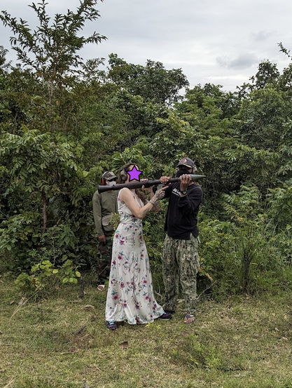 a woman in a flowy, flower pattern, floor length dress holds a rocket propelled grenade launcher, buffeted by a Cambodian soldier, as she is about to fire