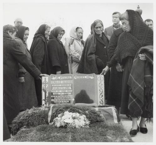 The image is a black and white photograph depicting a group of people gathered around a headstone. The headstone has text, which indicates it is for someone named Tenga de Amarangue who died on June 6th at the age of 45 years old. Two fur blankets are placed in front of the stone with flowers arranged atop them and beside them. Many individuals wearing dark coats or cloaks appear to be mourning; some wear headscarves, suggesting a possible cultural context related to grief rituals. The atmosphere seems solemn as people bow their heads and stand close together around the monument.