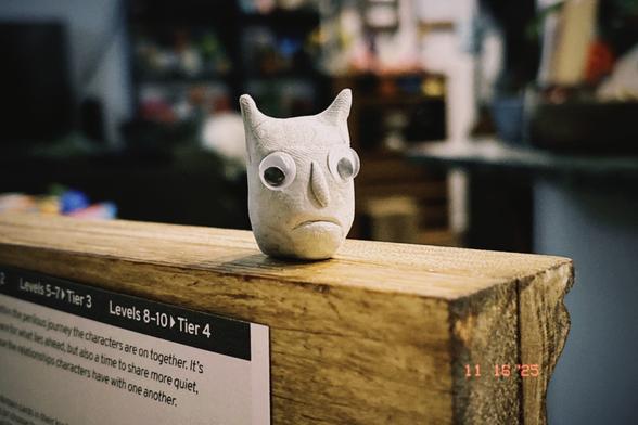 A little ball of Blu-Tack with googly eyes, a grumpy face, and horns. It is sitting on top of my wooden Game Master screen and looking a bit sad.