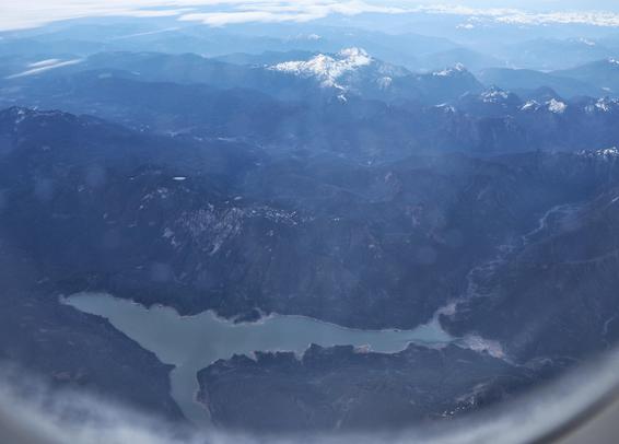 A stunning aerial view of a mountainous landscape, photographed from an aircraft window. Dominating the scene is a winding lake, its shape remarkably resembling that of a humpback whale from above. The lake’s calm, light blue waters contrast with the dense, shadowy valleys and forested hills that surround it. In the distance, towering peaks—some dusted with snow—rise against a hazy horizon, where layers of mist and clouds add a sense of depth and serenity to the scene.