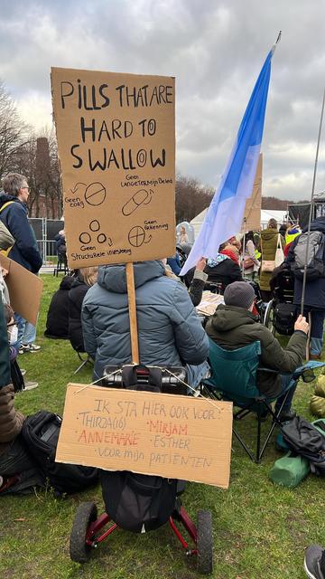 Demonstrant op de rug gezien in een hulpwagen met een tweetal kartonnen protestborden. Op de een staat "Pills that are hard to swallow" Daarnaast enkele tekeningen van verschillende soorten pillen met pijltjes en teksten: "Geen zorg", "Geen langetermijnbeleid", "Geen structurele investering", "Geen erkenning"