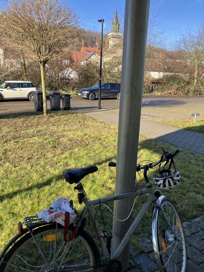 Ein altes graues Rad lehnt an einer Säule. Ein Helm hängt am Lenker. Dahinter eine sonnige Landschaft mit einer Kirche, im Vordergrund Mülltonnen, Gras und zwei Dosen.