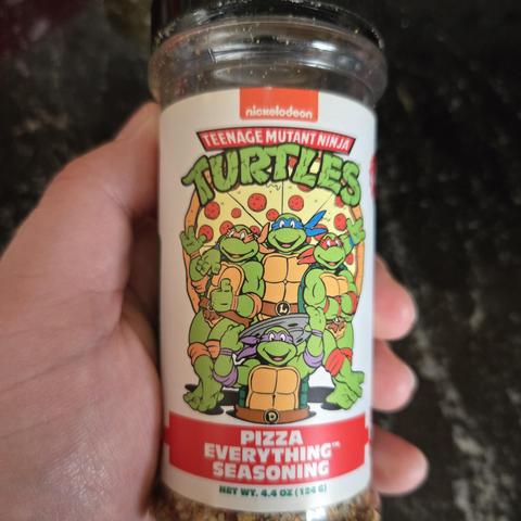 Photograph of a white person's hand holding a cannister of Pizza Everything Seasoning with the Teenage Mutant Ninja Turtles standing above the name of the product as well as their logo. They are drawn in their classic 1980s cartoon style.