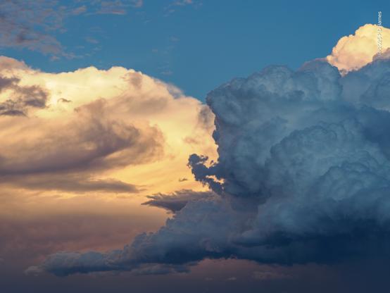 Two billowing cumulus storm clouds fill the frame, one brightly lit apricot by the setting sun, the other a shadowy blue grey. Above and behind them the sky is still blue.