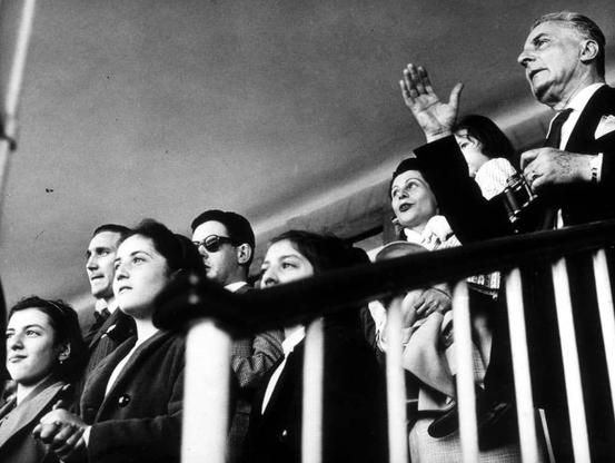 The image depicts a group of individuals seated closely together on what appears to be an elevated platform or balcony. The scene is rendered in black and white, suggesting it may have been taken several decades ago when color photography was not as prevalent.

In the foreground, there are multiple women; one woman stands out with her hand raised above her head, seemingly clapping or cheering enthusiastically. Another woman next to her appears to be holding binoculars, possibly observing something in the distance. The expressions on their faces range from anticipation and excitement to contemplation.

Behind them is a man who seems to be addressing someone off-camera; his right arm is extended upwards as if gesturing or pointing at something specific above him. His mouth is open, indicating he might be speaking loudly or expressing strong emotions.

The attire of the individuals suggests a formal event—men in suits and ties, women wearing dresses that suggest mid-20th-century fashion trends. The overall atmosphere conveys an intense moment shared among spectators engrossed by what they are watching from above them.

Additional information about this image can be found on Colombo Cesare's website through the provided URL: [https://images.loener.nl/RivistaFerrania/full/66f1/66f13b6c26a552dad254c350.jpg](https://images.loener.nl/RivistaFerrania/full/66f1/66f13b6c26a552dad254c350.jpg). This image appears to be part of a coll [...]