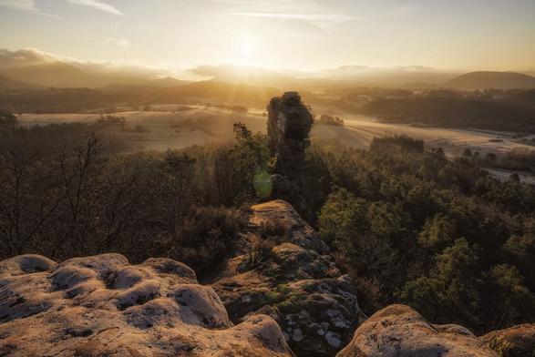 Zu sehen ist eine Sonnenaufgangsszenerie, bei der die Sonne im Bild die ganze Umgebung mit warmem Licht füllt. Selbst stehe ich auf einem großen Felsen und im Mittelgrund steht auch noch ein zweiter, von Bäumen umringter Fels.