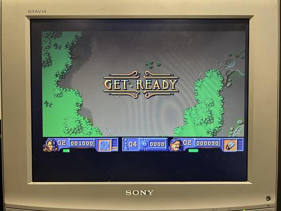 A photo of my silver Sony Bravia 4:3 flatscreen TV showing the Amiga game The Chaos Engine. The game is showing the start of the first level, which is a top down view of a grassy area with green trees around the playing area. Victorian era style text saying GET READY is at the centre of the screen.