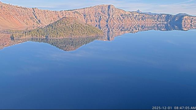 Blue sky, calm conditions, and an attractive reflection of Wizard Island and some of the geological features of Crater Lake National Park in this view from the Sinnott Memorial Overlook today. The blue color of the lake surface includes a few white cloud reflections as well. Vegetation on the island is green, while the surrounding slopes are orange, yellow, grey, and black. Mount Thielsen is indeed in view today. Look for the distant peak, upper right.
