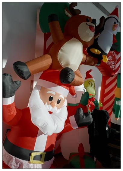 Large inflatable Christmas display featuring Santa Claus in the foreground with candy-cane-striped arms crossed. Behind Santa, several other festive inflatables including a reindeer, a penguin on a polar bear, and the Grinch, each decorated with holiday elements like scarves and hats. The setting appears indoors against a soft-lit wall.