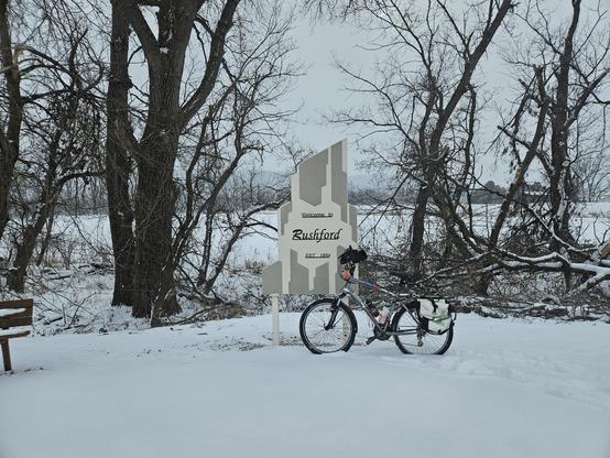 A grey bicycle with a white bag leans against a sign with an abstract white and grey design. In black letters "Welcome to Rushford Est. 1854". White snow is on the ground and the leafless trees under a grey sky.