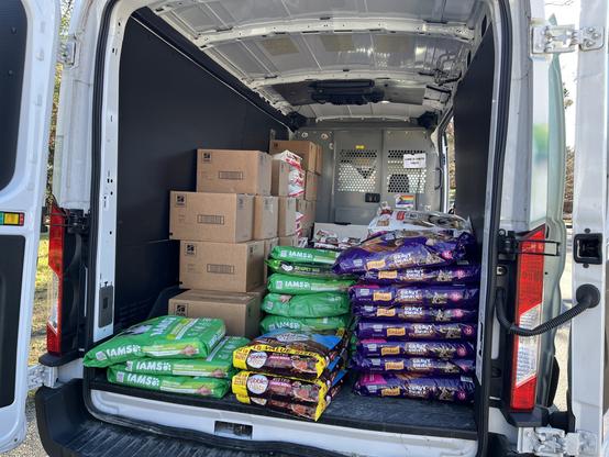 An mspca community outreach van packed with food for distribution. on the left, purple bags of friskies cat food are piled high. in the middle and on the right are green iams dog food bags as well as a few bags of kibbles and bits. there are also some cardboard boxes