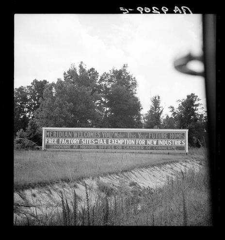 The image is a black and white photograph depicting an outdoor scene with a signboard prominently displayed in the foreground. The sign reads "MERIDIAN WELCOMES YOU / Make This Your FUTURE HOME FREE FACTORY SITES-TAX EXEMPTION FOR NEW INDUSTRIES INQUIRE AT MAYORS OFFICE OR CHAMBER OF COMMERCE." It is set against a backdrop of tall trees and an open field, with overcast skies above. The signboard's text invites potential businesses to Meridian by offering tax exemptions for new industries in exchange for locating at the mayor’s office or chamber of commerce.

Additional context: This photograph appears to be from Dorothea Lange's series capturing various aspects of life during a time when industrialization was transforming communities, particularly in Southern states. The image reflects an era where towns like Meridian were seeking ways to attract new businesses and industries for economic growth through incentives such as tax exemptions.

Further information: For more on the context behind this photograph or similar works by Dorothea Lange, it would be beneficial to explore her body of work during that period, particularly focusing on Southern industrialization. The image is part of an untitled collection which may provide additional historical and contextual insights into life in post-war America.