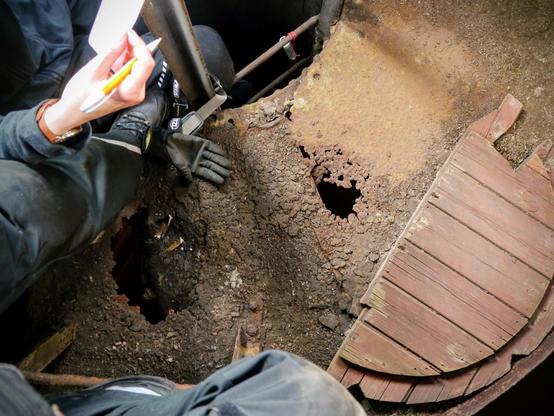 Foto im Innenraum eines alten Wasserturms. Der Fußboden ist rostig und mit zwei zackigen Rostlöchern. Rechts licht ein alter Holzdeckel. Eine Person sitzt in einer oberen Leiter, eine Person am Absatz auf dem Rand an einer unteren Leiter. An einer Spross baumelt angebunden ein PET-Preform. Eine Person hält einen entrollten Zettel und einen Kugelschreiber.