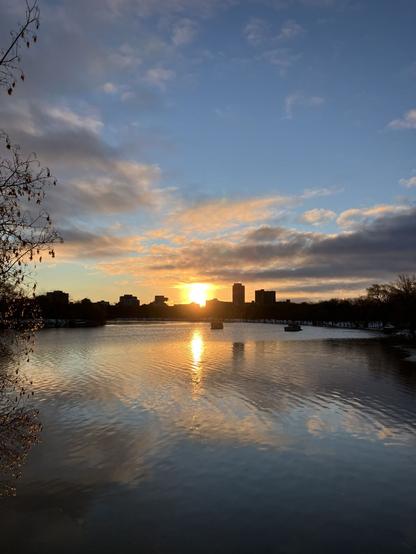 Bright sun beaming through clouds and blue sky rising behind cityscape skyline, reflecting on the partially frozen river.