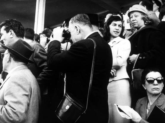 The image depicts a bustling public scene captured in black and white. In the foreground, various individuals are engaged with different activities; some appear to be taking photographs or making phone calls into large handheld devices characteristic of mid-20th-century technology.

A man is prominently featured in the center-left part of the frame, wearing a dark suit jacket over a lighter shirt, glasses, and holding what appears to be a professional camera. His posture suggests he's deeply focused on his task amidst the crowd around him.

Other people surround this central figure; some are standing close by with similar cameras or phones, indicating that they might also be photographers or reporters. There is noticeable diversity among the group: men in suits and hats, women wearing dresses and coats typical of mid-century fashion, all contributing to a sense of urgency or importance surrounding whatever event they're covering.

The background shows more people in various states—some looking directly at the camera with expressions ranging from curiosity to mild concern. The lighting is stark, creating strong contrasts between light and shadow on their faces and clothing, which adds drama to the scene.

Overall, this image captures a moment that seems to be of significant public interest during what could possibly be an important political event or social news happening at the time as indicated by its black-and-white style.