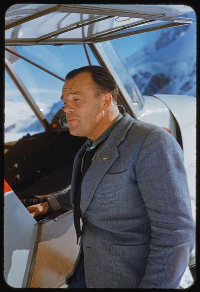 A man in a grey wool coat standing beside the cockpit of an aircraft with its canopy open; snowy mountains are visible through it. This photograph is from Toni Frissell's collection, dating between 1930s and 1950s, featuring subjects like aviation, skiing, travel, and Switzerland. The specific location for this image was Zermatt in February of the year 1955.