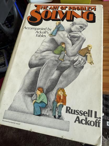 A book cover titled "The Art of Problem Solving" by Russell L. Ackoff features a large, thoughtful statue figure with four smaller illustrated characters sitting on it. The cover includes the subtitle "Accompanied by Ackoff's Fables"