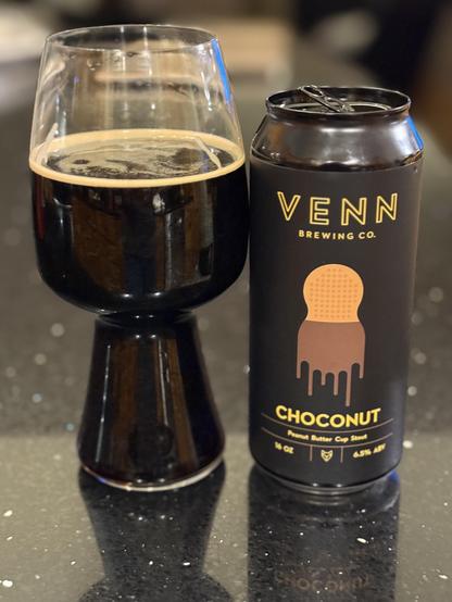 A stout glass full of dark beer. The black pint can is to the right and has yellow lettering and a chocolate dipped peanut shaped cookie