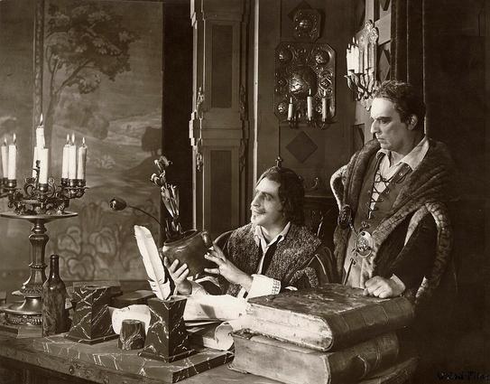 Eduard Verkade and Dio Huysmans as the de Witt Brothers. Black and white.
The photograph, rendered in sepia tones, features a detailed scene set with a man seated at a desk, engrossed in inspecting a vase with flowers and a quill inside. To the left, a tall candelabra holds lit candles, casting a warm glow. On the desk, various marble objects, a bottle, and scattered papers suggest a workspace filled with creative activity. To the right, another man stands beside the desk, wearing a decorative cloak and a serious expression. He rests one hand on a stack of large, aged books that sit on the edge of the desk.