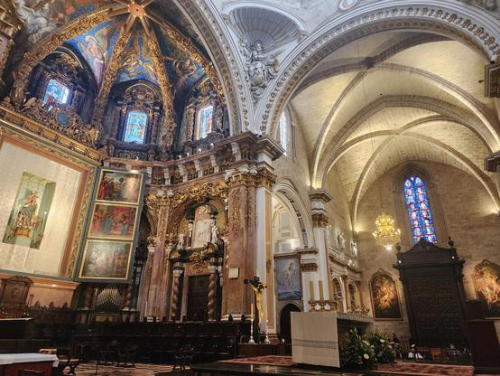 Wide-angle shot of, at left, apse with opulent gold relief, monumental altarpiece with scenes from the life of the virgin and a statue of her at center. above are stained glass windows and frescoes of singing angels in a da Vinci-like style. To the right is a direct view of one arm of the cathedral, with vaulted ceiling, large wooden closet(?), tall narrow stained glass window, chandelier, and paintings of religious scenes on either side of the closet.