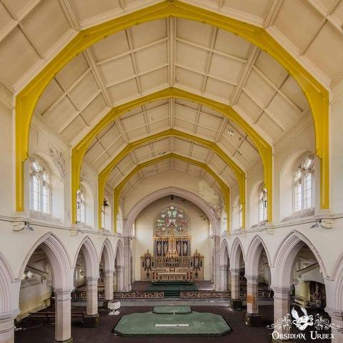 Interior of a large, empty church with vaulted yellow and white ceiling, arched columns, stained glass windows, and an ornate altar at the far end.