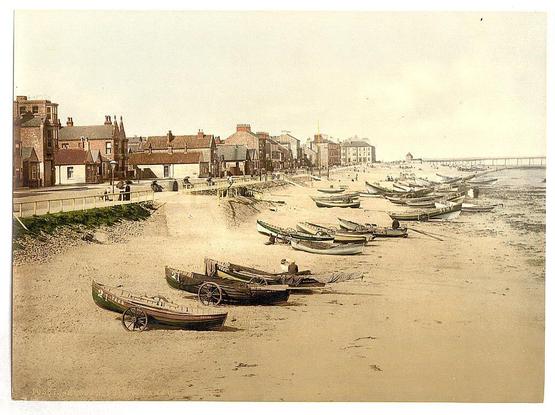 The image depicts a coastal scene with numerous boats lined up along the shore. These wooden vessels appear to be fishing or rowboats, and some are equipped with masts suggesting they could also function as sailing crafts when conditions permit. The beach is sandy and there's evidence of recent use; footprints and tire tracks crisscross the sand, indicating human activity.

In the background, a series of quaint buildings form what seems to be an esplanade or seaside promenade typical for towns in England during that era. These structures vary from simple cottages with gabled roofs to more substantial brick buildings, possibly serving as shops or residences. A row of lampposts runs along part of the path, providing light.

The sea extends into the distance where a bridge can be faintly discerned connecting two parts of land over water. The sky is clear and bright, suggesting good weather conditions during the time this photograph was taken.

Notable features include the vintage aesthetic suggested by the image's quality and coloration—hints of sepia tones that are characteristic of early photographs developed with photochrome or a similar process in the late 19th century to early 20th century. The absence of modern elements, like cars or contemporary clothing styles on any visible figures, supports this dating framework.

The photograph is credited as being from Redcar's esplanade in Yorkshire, England and was captured by [...]