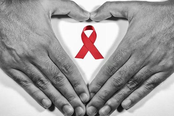 Two hands form a heart shape around a bright red HIV/AIDS awareness ribbon on a white background. The hands and background are in black and white, making the red ribbon stand out prominently.