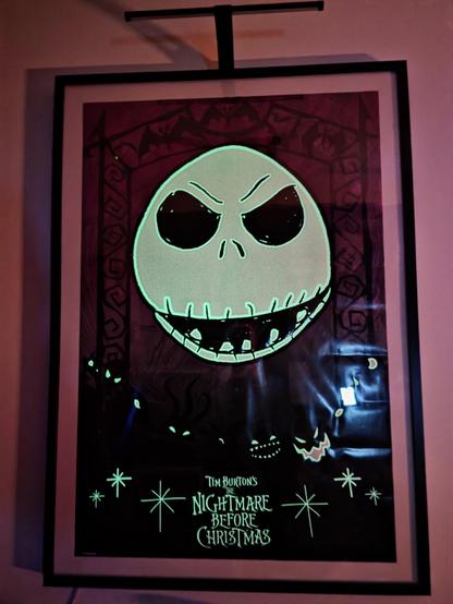 A framed glow-in-the-dark poster from Tim Burton’s The Nightmare Before Christmas hangs on a softly illuminated wall. At the center glows Jack Skellington’s iconic face: a large, eerie green skull with hollow black eyes, thin nostrils, and a jagged stitched grin that seems to float in the darkness. Behind him, faint purplish-red tones from the room’s candlelight give the poster a warm, haunting ambiance, subtly revealing background shapes like bats, twisted trees, and ghostly silhouettes from Halloween Town. Lower in the frame, glowing pumpkin faces and stylized stars surround the film’s title in bright green lettering. A bright white reflection on the right side—the unavoidable glare from the user’s weather station across the room—cuts through part of the poster’s surface, adding an unplanned but harmless highlight. The black frame outlines the artwork sharply against the dim surroundings.