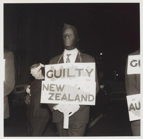 The black and white photograph depicts a man standing at night, holding up a sign that reads "GUILTY NEW ZEALAND." He appears solemn with his head slightly bowed. The text on the image is bold and clear against the contrasting background of darkness, emphasizing its message. Behind him are indistinct figures also bearing signs related to activism or protest. Some people seem to be looking towards something outside of the frame, possibly capturing this moment for documentation. There's a notable contrast between light and shadow in the photo which adds an element of drama to it. The image is dated from "National mobilisation against Vietnam war demonstration" as indicated by the caption below with reference number 6690f4db3c8dcd5ca3a70d4c.jpg, implying that this was taken during a significant political event in New Zealand's history involving anti-Vietnam War demonstrations. The image is credited to Ans Westra and appears as part of his photographic record on the website images.loener.nl.