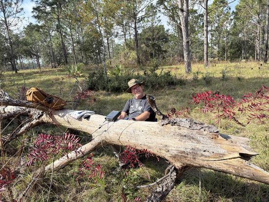 A man (me) sitting in a camp chair, behind a fallen log in a forest. The fallen log is being used as a table. All my radio gear and backpack are on the log.