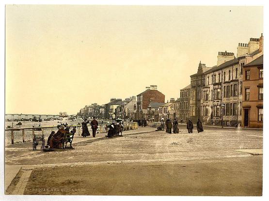 The image depicts a bustling street scene in an urban setting during what appears to be the late 19th or early 20th century. The photograph is sepia-toned, which suggests it could have been taken with photographic processes from that era.

In the foreground, there are several individuals walking along the pavement and others seated on benches, engaged in various activities such as conversing, reading newspapers, or simply resting. Many of these figures wear long coats indicative of a cooler climate typical for much of England during this period.

The street is lined with buildings displaying architectural styles characteristic of that time—stately rows of terraced houses with multiple stories and large windows, suggesting residential use. The structures exhibit varying facades in colors such as beige, brown, and muted reds or greens, common to the era's construction materials like brick and stucco.

In the middle ground, a group of people is gathered near what appears to be an entrance gate with railings leading into another part of this area. Their attire suggests they might be travelers or workers heading towards their destinations within the town. The street itself has minimal vehicular traffic but does accommodate horses as evidenced by at least one horse-drawn carriage.

The background showcases more architectural diversity, including a red-brick building and other structures with different designs that create an at [...]