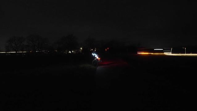 Bunt beleuchtetes Fahrrad auf einem Feldweg in der Nacht. Im Hintergrund verursacht ein Beleuchteter LKW Lichtspuren