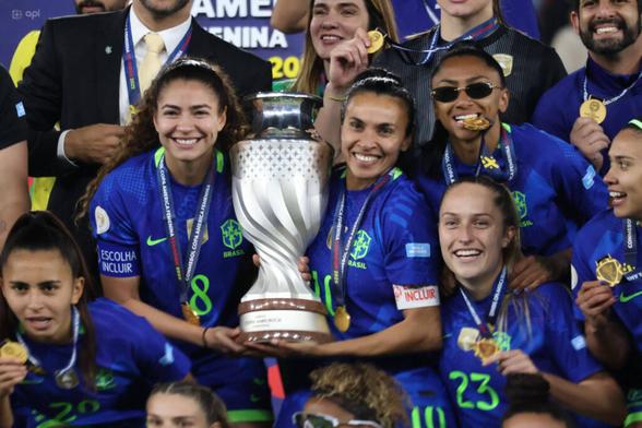 Brasiliens Spielerinnen mit der Trophäe der Copa América, sie tragen Goldmedaillen um den Hals und in der Mitte halten Angelina und Marta zusammen den silbernen Pokal. Alle um sie herum grinsen, manche haben Sonnenbrillen auf.
