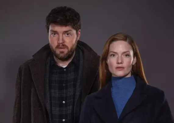 Zwei Personen stehen vor einem grauen Hintergrund. Der Mann trägt einen Mantel, die Frau einen blauen Pullover. Beide blicken ernst in die Kamera.