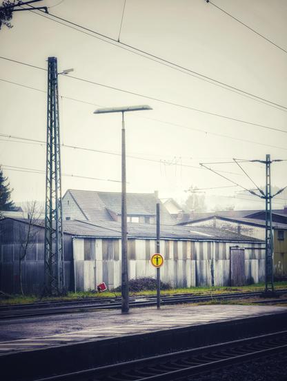 Hinter Gleisen und Bahnsteig liegen trüb Lagerhallen und Dächer.