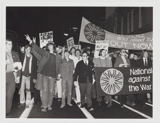 The photograph depicts a group of individuals participating in what appears to be an anti-war protest. It is taken at night, as indicated by the darkness surrounding them and artificial lighting illuminating their presence on the road. The main subjects are holding up signs with various slogans such as "National mobilisation against Vietnam war," which suggests they are part of a demonstration opposing U.S. involvement in the Vietnam War.

Several individuals carry flags or banners; one prominent banner reads, “All troops out of Indonesia now,” and another shows text that includes references to Australia (AUS), New Zealand (NZ), and an allied force. The attire varies among participants, with some wearing coats indicative of cooler weather conditions. A person is seen holding up a peace sign while others are gesturing in support.

One individual on the right side holds a distinctive banner featuring what looks like a stylized sun or starburst pattern within concentric circles, possibly representing an emblem related to their cause. The photo's monochromatic tone emphasizes its historical context and seriousness of purpose.