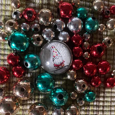 Ein Druckknopf wurde mit einem Wichtel bemalt. Der Wichtel hat einen langen Bart und trägt rote Kleidung und eine hohe weiße Mütze mit roten Punkten, die seine Augen verdeckt. Der Druckknopf liegt zwischen roten, grünen, silbernen und goldenen Perlen in unterschiedlichen Größen.