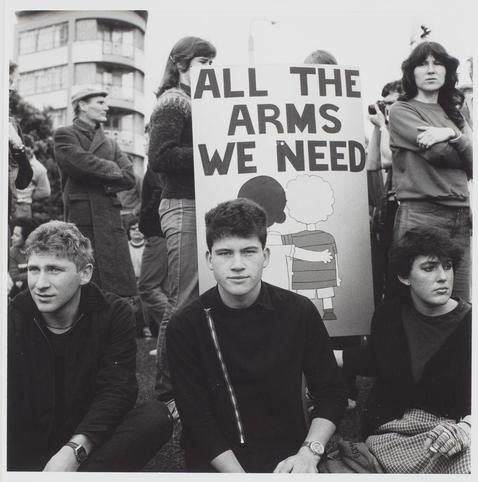 The black and white photograph captures a moment from an anti-nuclear weapons protest. In the foreground, three young individuals are seated in front of a sign with bold lettering that reads "ALL THE ARMS WE NEED," accompanied by simple illustrations depicting two figures—one child-like figure and one adult—which may symbolize innocence versus authority or necessity for arms use. The central individual appears contemplative while looking off to the side; their peers flank them, all dressed in casual attire typical of a protest scenario from an era where black jackets were prevalent. Behind them stands another person with folded arms, possibly conveying solidarity or defensive stance towards the message on display.

The background is filled with indistinct figures and urban architecture, suggesting that this gathering takes place outdoors within a city setting. The crowd's composition indicates diverse ages and genders among its participants, hinting at widespread public support for the cause against nuclear weapons in the context of Wakefield Street, as referenced by the source link provided.
