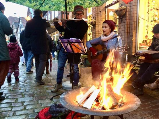 Straßenmusiker spielen Gitarre und Flöte vor einem Feuerkorb auf einem gepflasterten Platz. Im Hintergrund Menschen und Gebäude.