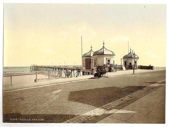 The image displays an old sepia-toned photograph depicting a coastal scene, likely from the late 19th to early 20th century. It shows Redcar Pier in Yorkshire, England during this period. The pier is seen extending into calm waters under a hazy sky with clouds suggesting it might be midday or afternoon due to brightness and lack of long shadows.

There are several structures on the right side of the image; two prominent domed-roof pavilions stand out, likely serving as refreshment kiosks. Between these buildings is a lamppost-like structure holding what appears to be an ornate light fixture. In front of these structures, there's a group of people seated at benches and on chairs under open umbrellas or canopies for shade.

The foreground features a paved sidewalk leading towards the pier where more individuals are seen strolling along it. The sand is visible alongside this path with gentle waves lapping against what could be the edge of an extension to the beach area not shown in full view due to perspective. A wooden railing separates the promenade from the sea.

The bottom left corner includes text stating "103-85 REDCAR, THE PIER," which likely refers to a cataloging or reference number for this specific image within a collection. The overall atmosphere of the photograph conveys leisure and relaxation typical of seaside resorts during that era in Britain.