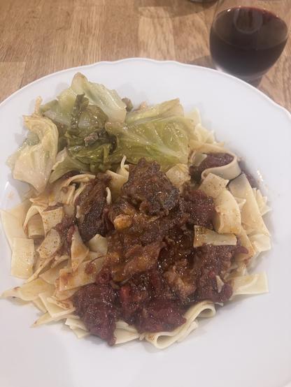 A plate of noodles topped with a hearty meat sauce and accompanied by cooked green cabbage. A glass of red wine is visible in the background.