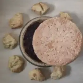 Se démarquant sur le fond d'une table beige, le dos d'un pain rond roussi masque partiellement le disque d'une tarte aux myrtilles sur fond de purée d'abricots. Sept choux, disposés en arc de cercle, ornent la partie visible de la tarte. Dans le croissant qui reste apparent, les myrtilles cuites constituent un fond sombre laissant apparaître quelques rares tâches oranges. Le disque de la tarte est serti du beige clair du fond de tarte, puis du verre transparent du plat en Pyrex. Les choux au fromage sont jaunes un peu roussis.
