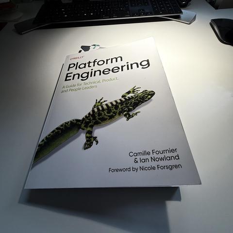A photo of the O’Reilly book “Platform Engineering: A Guide for Technical, Product, and People Leaders” lying on a white desk. The book’s cover features a large illustration of a green-and-black salamander. A keyboard, mouse, and other desk items are slightly visible in the background