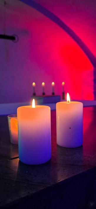 Zwei brennende weiße Kerzen im Vordergrund, mehrere Kerzen im Hintergrund. Zu sehen ist außerdem ein Gewölbedecke, die in rötliches Licht getaucht ist.
