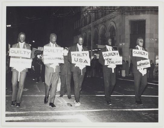 The image depicts a nighttime street scene where five men are walking side by side, each holding up signs with the word "Guilty" followed by different country names. The countries listed include South Korea, Philippines, USA, New Zealand, and Australia. They appear to be participating in some form of protest or demonstration. All individuals wear dark suits and ties except for one man on the far right who is dressed more casually in a suit with an open-collar shirt. Their expressions are serious as they march forward together. The background reveals part of an ornate building, suggesting this event takes place near significant architecture possibly indicative of government buildings or institutions. The overall tone suggests a historical context likely related to political activism against Vietnam during the 20th century era commonly known for anti-war movements.